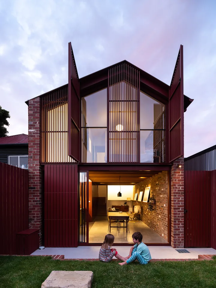 A narrow two-storey brick home with dark red shutters folded open at dusk, its rooms lit, two people sitting on the lawn outside